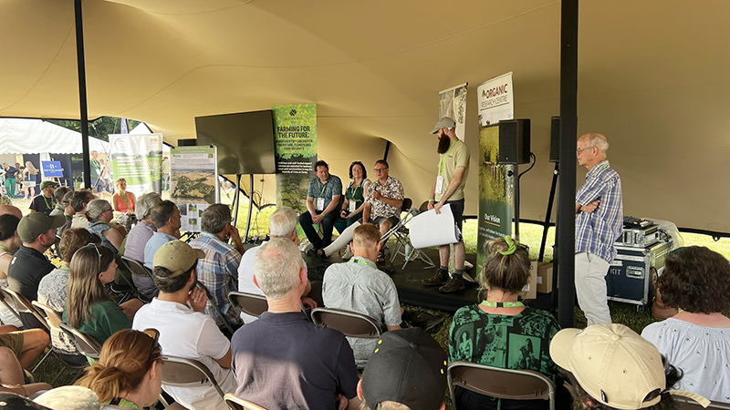 A tent full of people listening to presenters on the stage talking about Wakelyns and its legacy