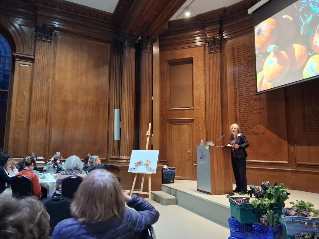 Kath Dalmeny on platform  in panelled room addressing a seated audience, with crates of vegetables on one side and a slide of vegetables on the screen.