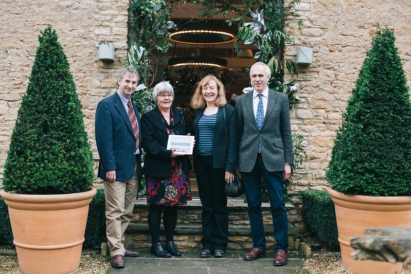 Four people stand outside a doorway framed by potted trees