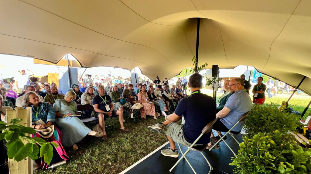 View of agroforestry tent from behind stage with speakers backs and audience facing the camera